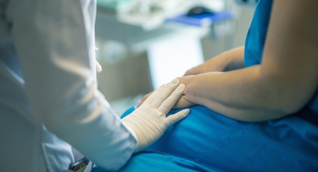 En klinisk bild som visar en vårdgivare som bär medicinska handskar och håller en patients hand i vad som verkar vara en medicinsk vårdmiljö. Detta representerar medkännande hälsovård och relationen mellan vårdgivare och patient.