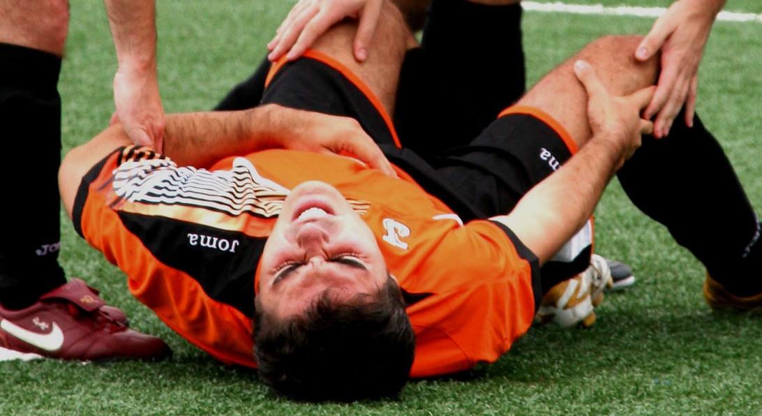 En actionfotografi som visar medicinsk personal som tar hand om en skadad idrottare i en orange sportuniform som ligger på en spelplan. Detta skildrar den omedelbara responsen på en idrottsskada och belyser vikten av medicinskt stöd på fältet som en del av idrottens hälso- och sjukvårdssystem för förebyggande och hantering.